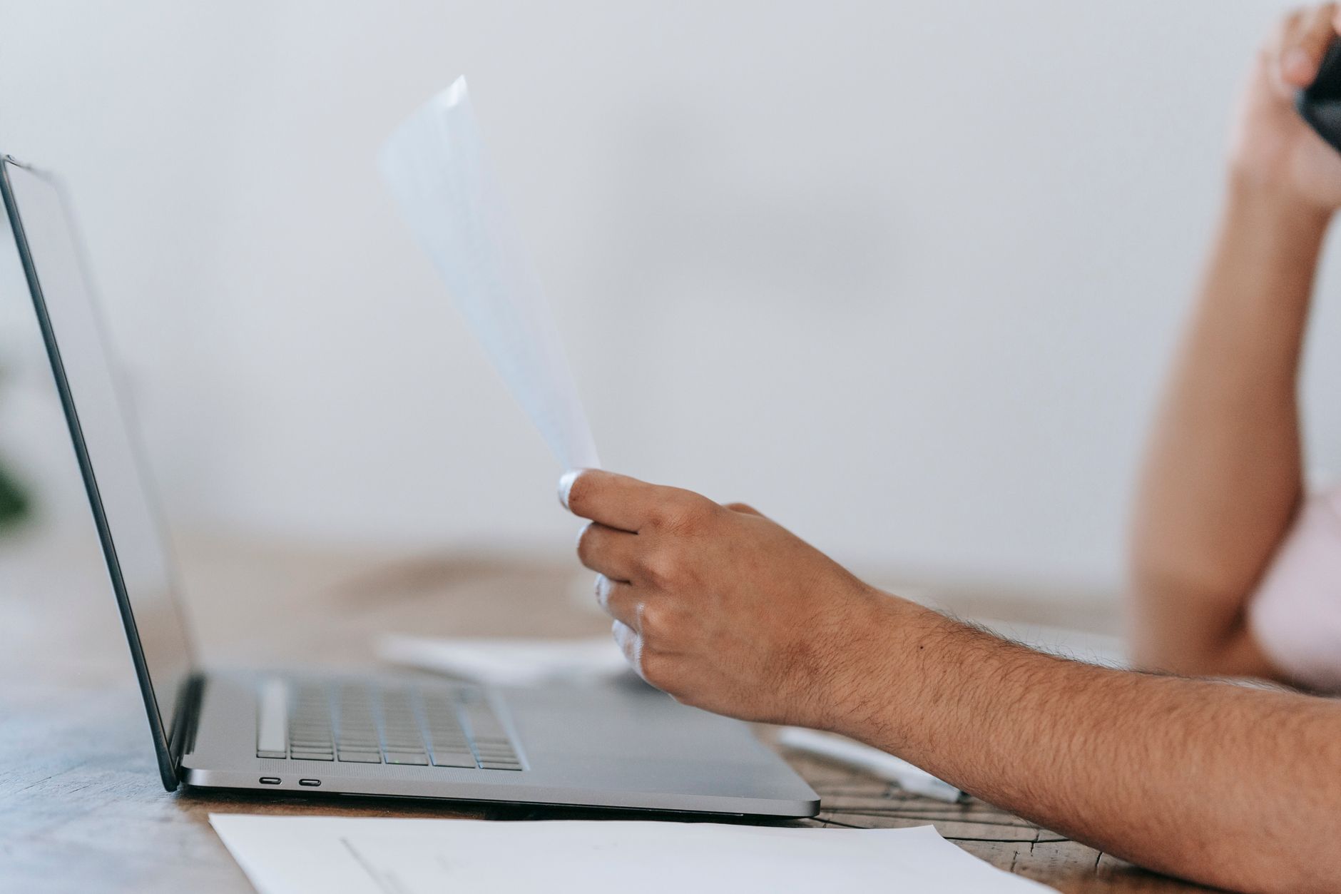 Homme anonyme recadré lisant un document et parlant au téléphone portable tout en étant assis à un bureau avec un ordinateur portable.