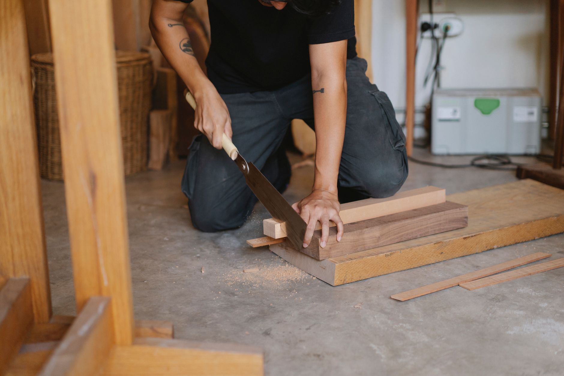 Artisan agenouillé dans un atelier qui scie une pièce de bois avec une scie à main, des planches et de la sciure au sol.