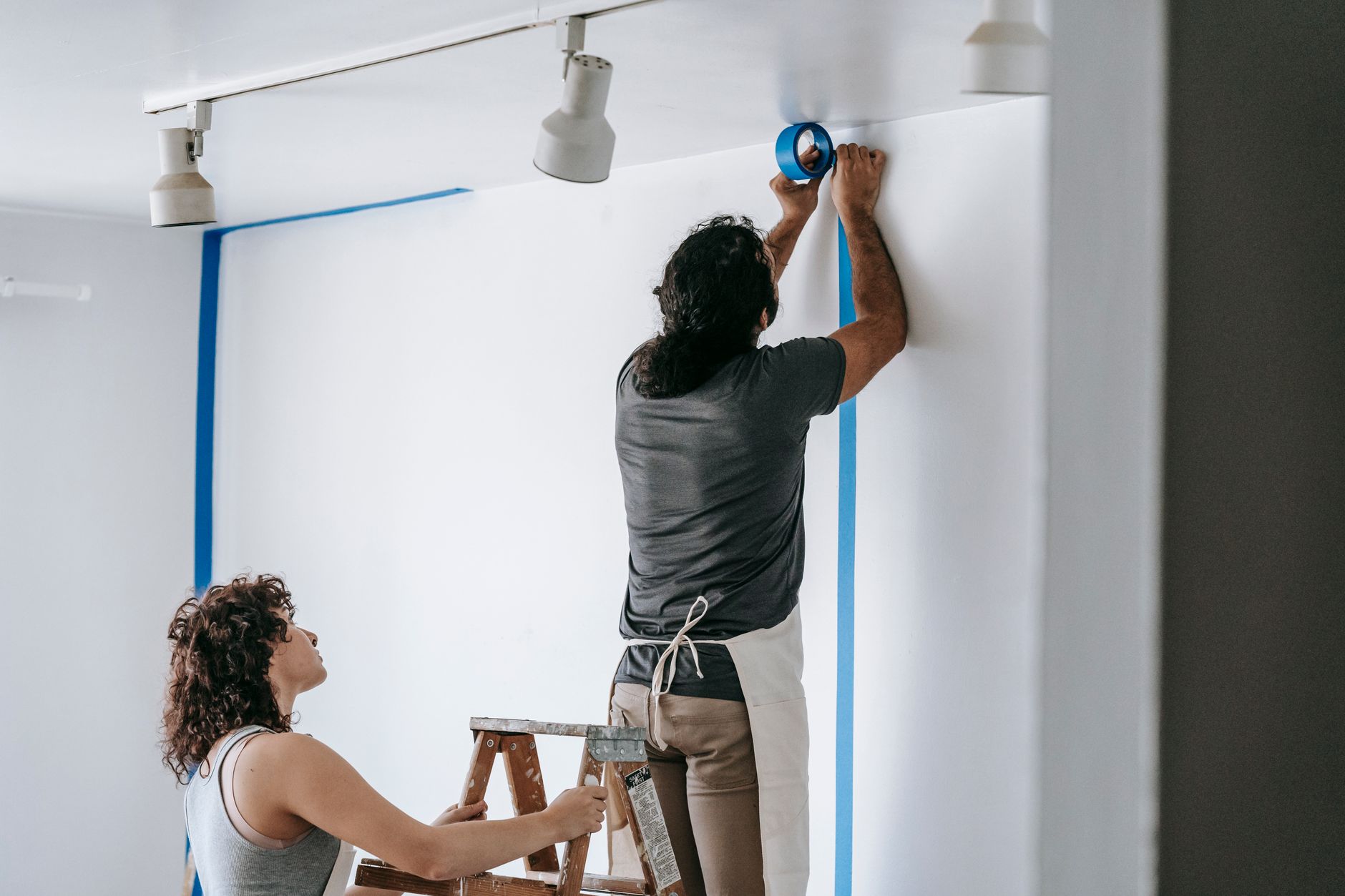 Deux personnes préparent un mur blanc : l’une, sur un escabeau, pose du ruban de masquage bleu, l’autre tient l’escabeau.